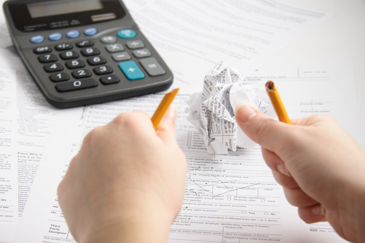 A person feeling overwhelmed while doing taxes crumples up a receipt in frustration, with a calculator, pencil, and tax documents spread out on a table.