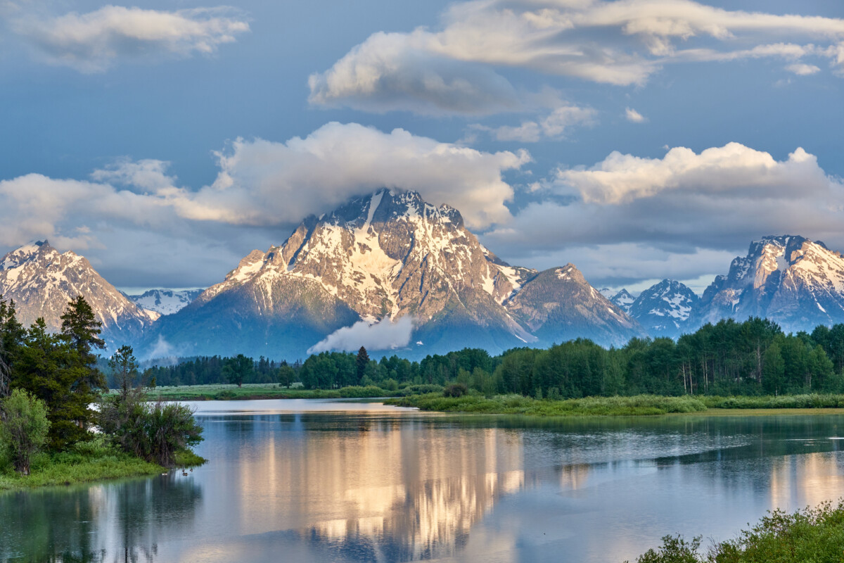 Majestic mountain peaks crowned with snow, reflecting on the tranquil waters of a serene lake under a softly lit sky with hints of blue and scattered clouds.