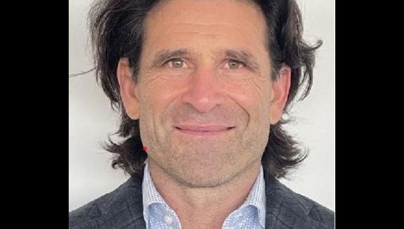 A person with shoulder-length dark hair is smiling at the camera. They are wearing a dark blazer over a light blue shirt. The background is a plain light color.