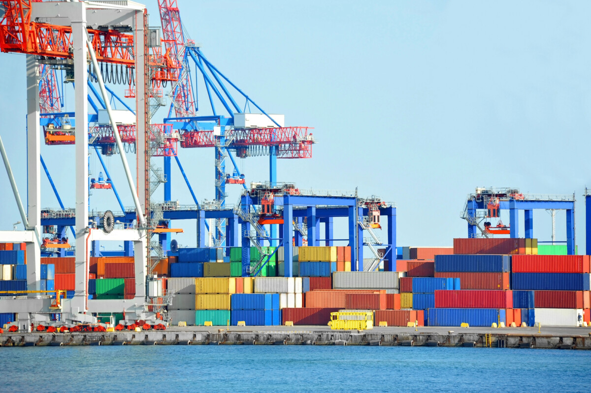 Colorful shipping containers are stacked at a busy port, with large blue and red cranes above them, ready for loading or unloading cargo; the scene is set beside calm blue water under a clear sky.