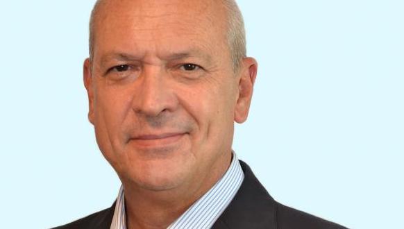 A middle-aged man with short gray hair, wearing a dark suit jacket and a light blue striped shirt, is smiling slightly while looking at the camera against a plain light background.