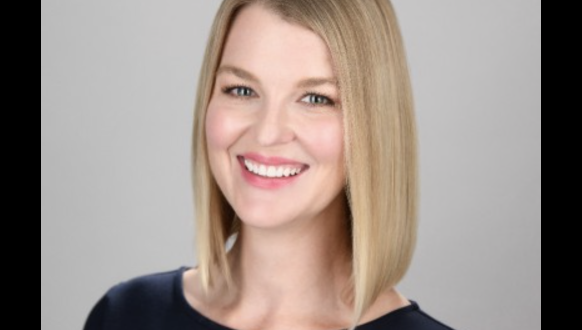 A woman with straight, shoulder-length blonde hair and a side part smiles at the camera. She is wearing a navy blue top and is posed in front of a plain, light gray background.