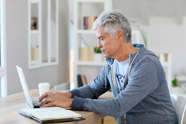 A middle-aged man with gray hair wearing a hoodie is sitting at a desk, working on a laptop. There is a notebook and a coffee cup on the table, with bookshelves in the background.