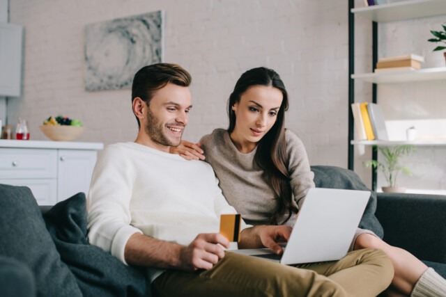 A smiling couple sits on a couch at home, looking at a laptop. One holds a credit card, suggesting they are shopping or browsing online together in a cozy living room setting.