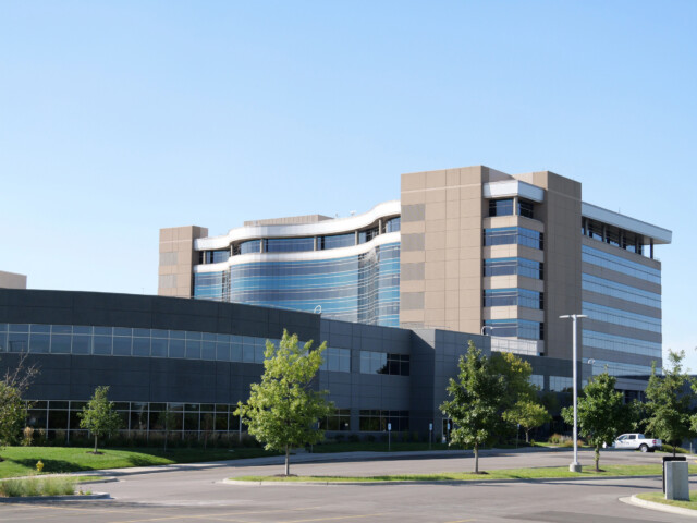 A modern building - Garmin HQ - with large windows, multiple stories, and a curved glass section, surrounded by green grass, trees, and an empty parking lot under a clear blue sky.