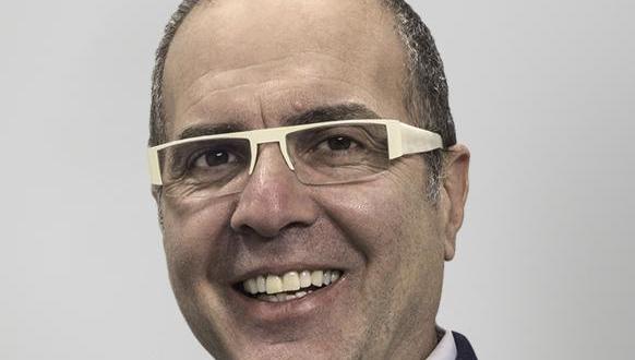 A middle-aged man with short hair and glasses smiling, wearing a suit jacket, white shirt, and tie, against a plain light gray background.