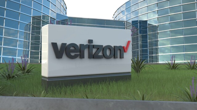 A large Verizon sign stands on a grassy area in front of modern glass office buildings with purple flowers and plants nearby.