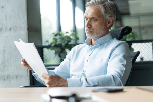 A middle-aged man with gray hair sits at a desk in an office, wearing a light blue shirt and reading documents with a thoughtful expression. There are plants and large windows in the background.